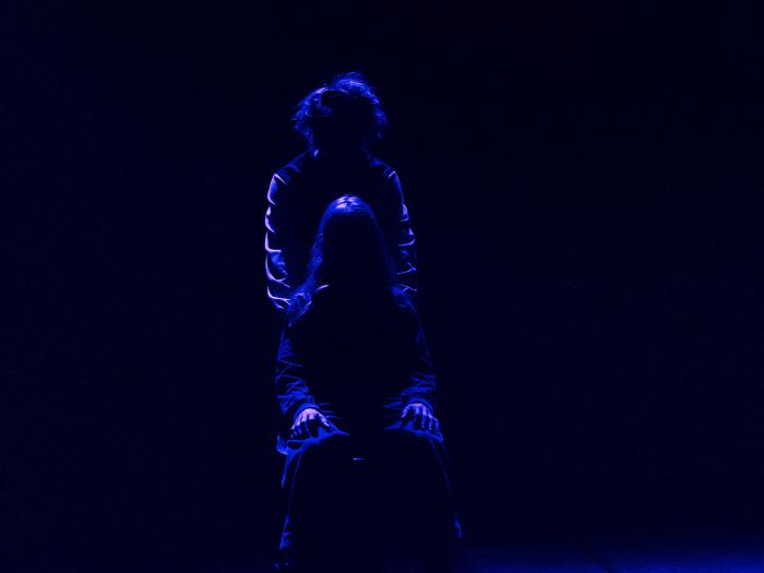 Mørk scene med blåt baglys. To skikkelser, en siddende og den anden bagved